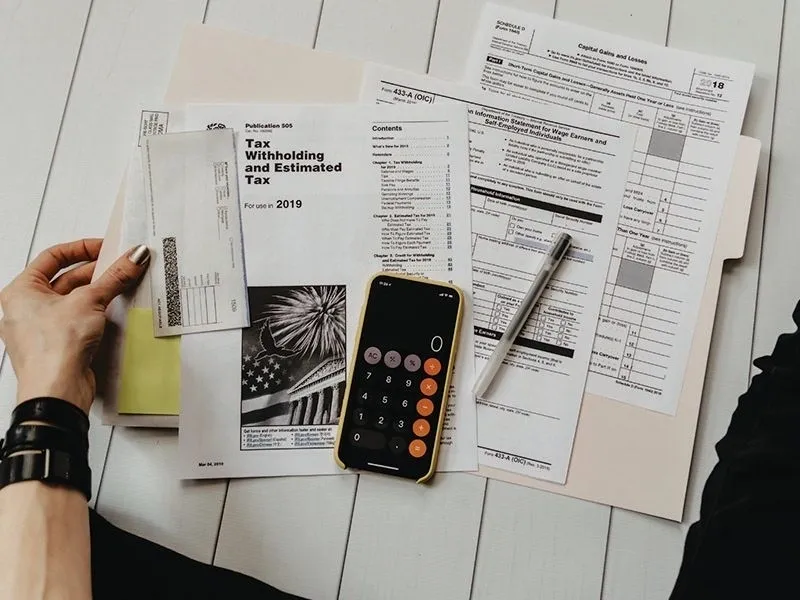 A person 's hand holding an open calculator next to some papers.