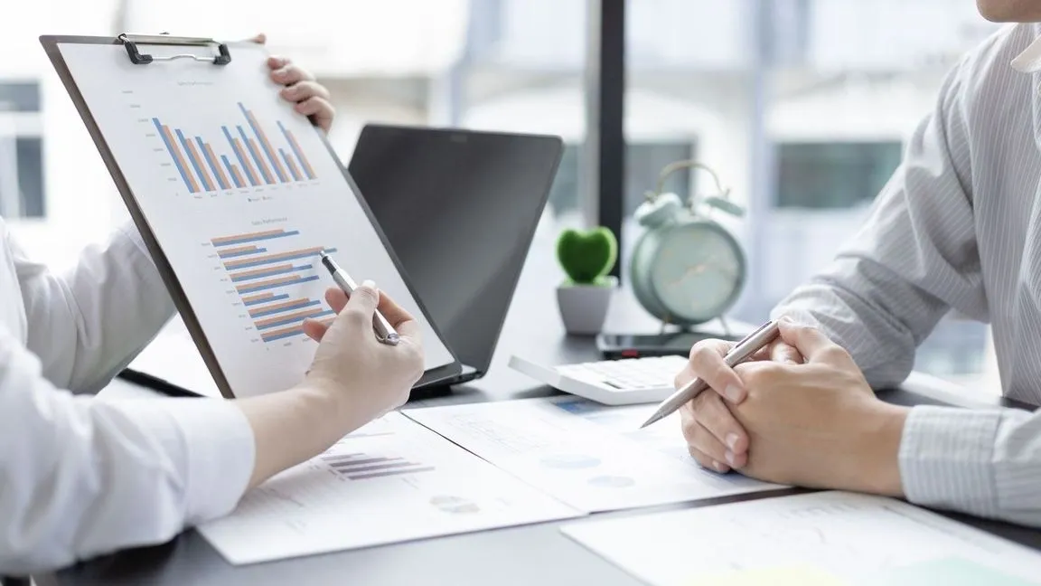 Two people are sitting at a table with papers and pens.