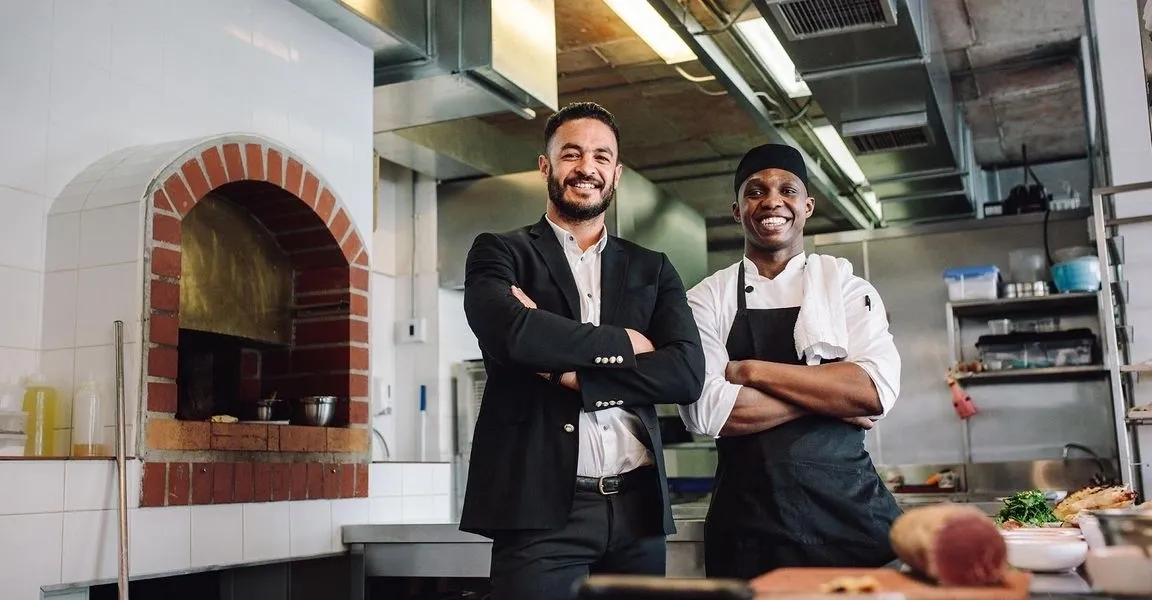 Two men standing in a kitchen with their arms crossed.